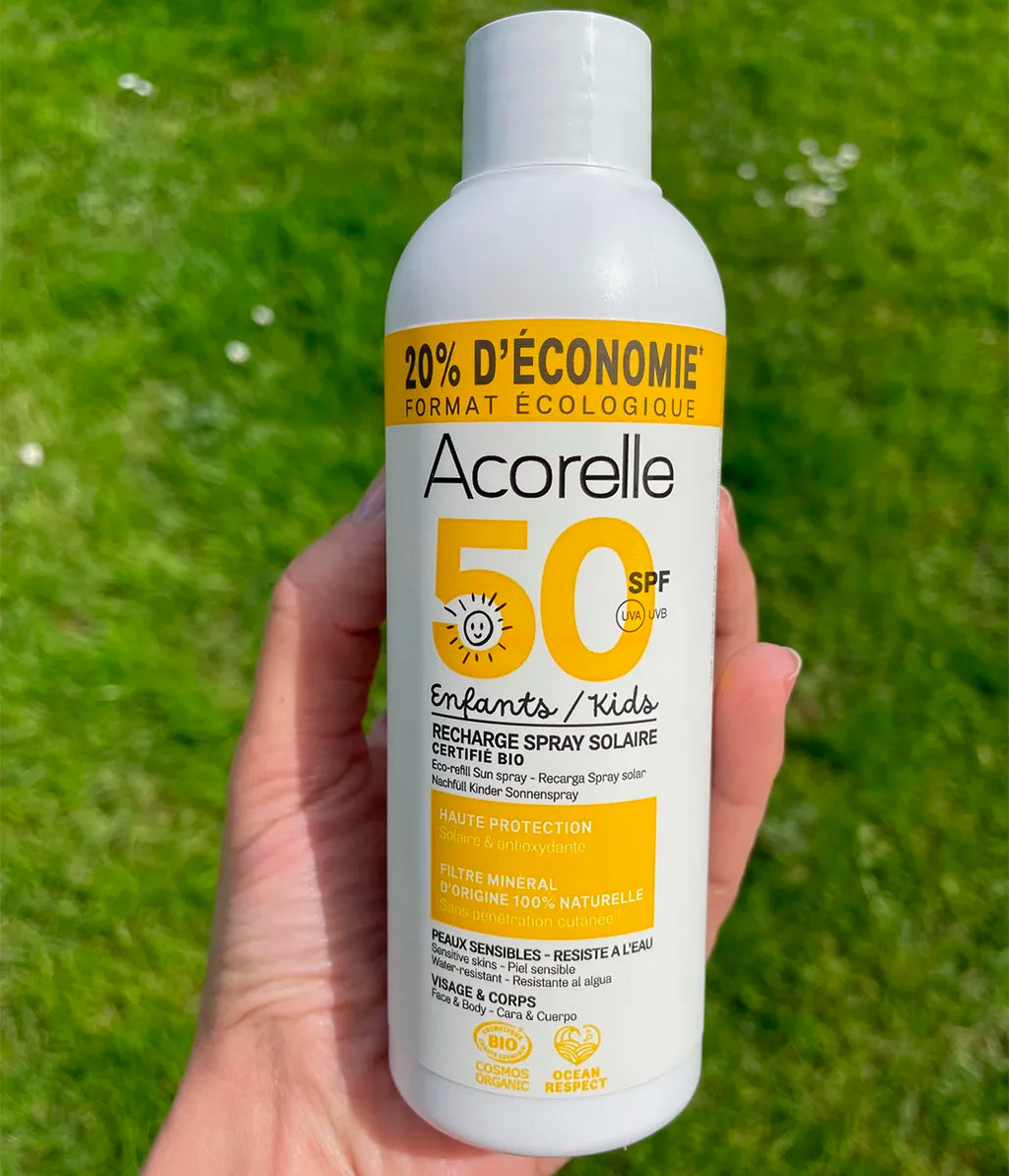 Mão a segurar a embalagem de recarga do protetor solar Acorelle SPF50+ para crianças num relvado.