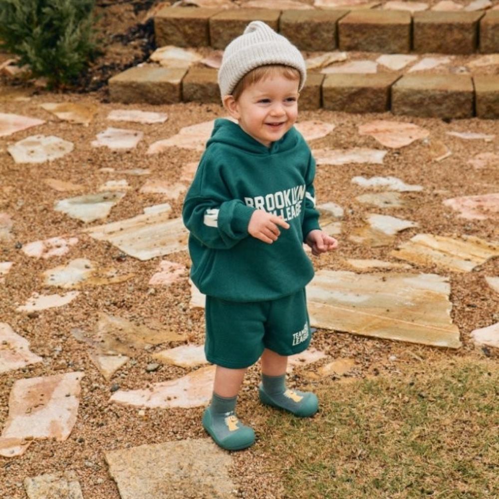 Bebé sorridente, vestido com um conjunto verde e gorro cinzento, a usar sapatos Attipas DINO GREEN, de pé num jardim.