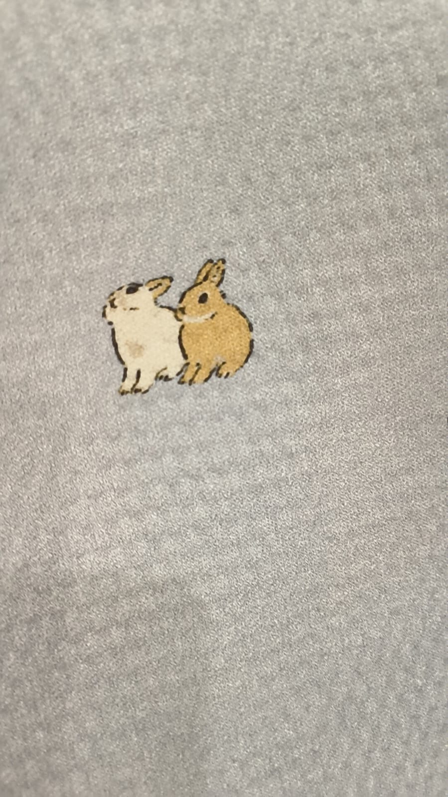 Detalhe em close-up do tecido cinzento com um pequeno bordado ou estampa de dois coelhos juntos (um branco e um castanho).