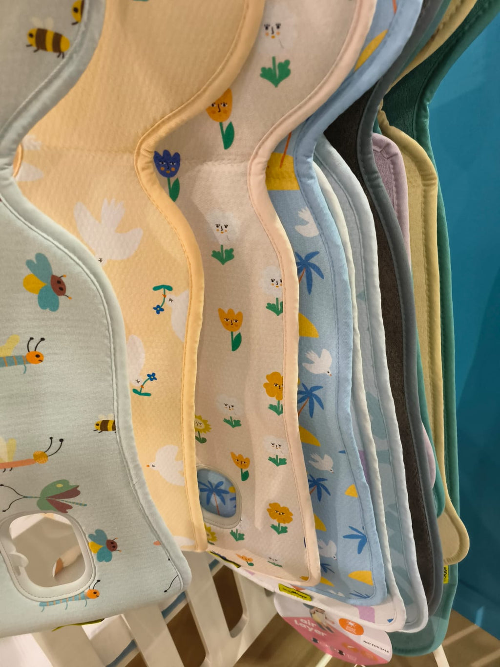 Várias capas AirLayer 3D Aeromoov penduradas, exibindo uma variedade de cores e padrões infantis (flores, insetos, pássaros).