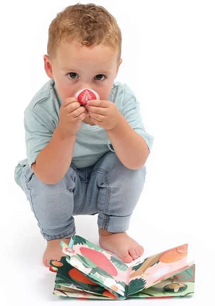 Criança pequena a cheirar uma das peças de morango do Ludi Livro de cheiros - Frutas, demonstrando a interação sensorial.