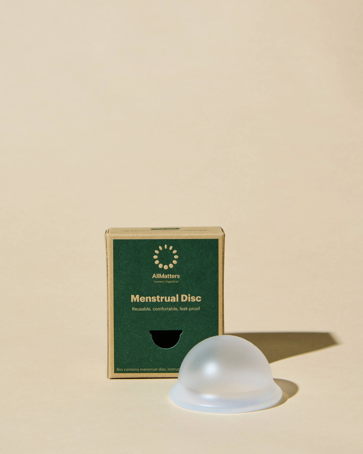 O Disco Menstrual AllMatters translúcido posicionado ao lado da sua caixa de cartão verde, num cenário neutro e iluminado.