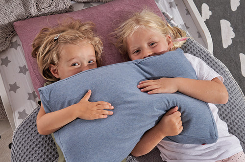 Duas crianças loiras a sorrir, deitadas lado a lado, abraçando uma almofada Theraline azul, em cima de uma cama com cobertores.