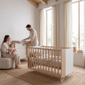 Micuna Amaia Berço 120x60 em Madeira / Branco num quarto de bebé luminoso, com os pais e o bebé ao lado do berço.