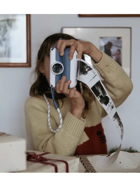 Criança a usar a máquina fotográfica KidyWolf K-PRINT azul e branca, tirando uma foto que é impressa em formato de tira contínua em preto e branco.