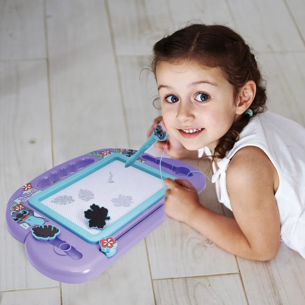 Criança sorridente deitada no chão a usar o quadro magnético roxo do Stitch para desenhar com a caneta de água.