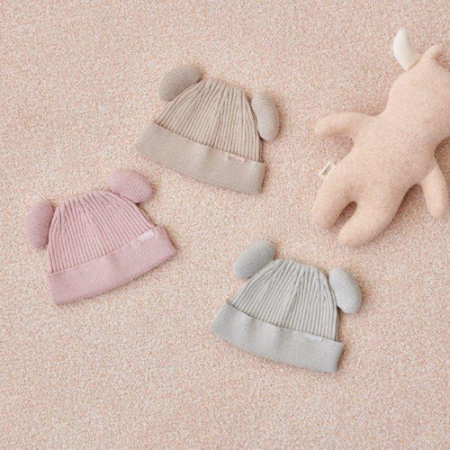 Três gorros de malha Attipas com orelhas em tons de rosa, bege e cinza, dispostos sobre um fundo texturado rosa, junto a um peluche.