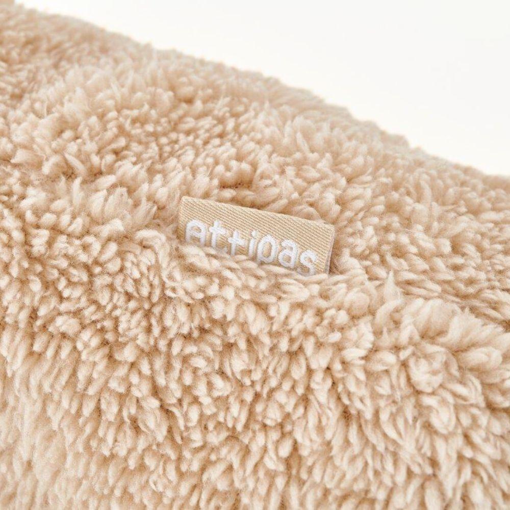 Detalhe em close-up da textura fofa e da etiqueta com o logótipo "attipas" num aquecedor de pés bege.