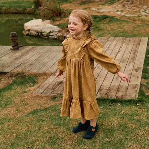 Criança sorridente com cabelo apanhado, vestindo um vestido mostarda, a usar os sapatos Attipas Skin Shoes verde escuro num ambiente exterior com relva e um lago.