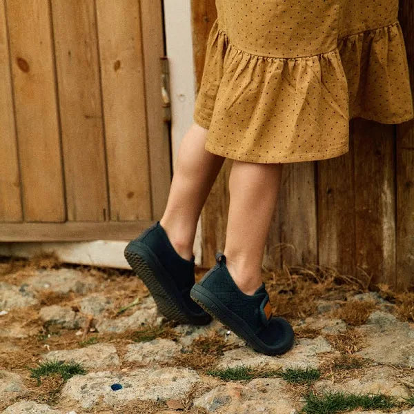 Detalhe dos pés de uma criança a usar os sapatos Attipas Skin Shoes verde escuro, junto a uma parede de madeira e em terreno irregular, destacando a sola flexível.