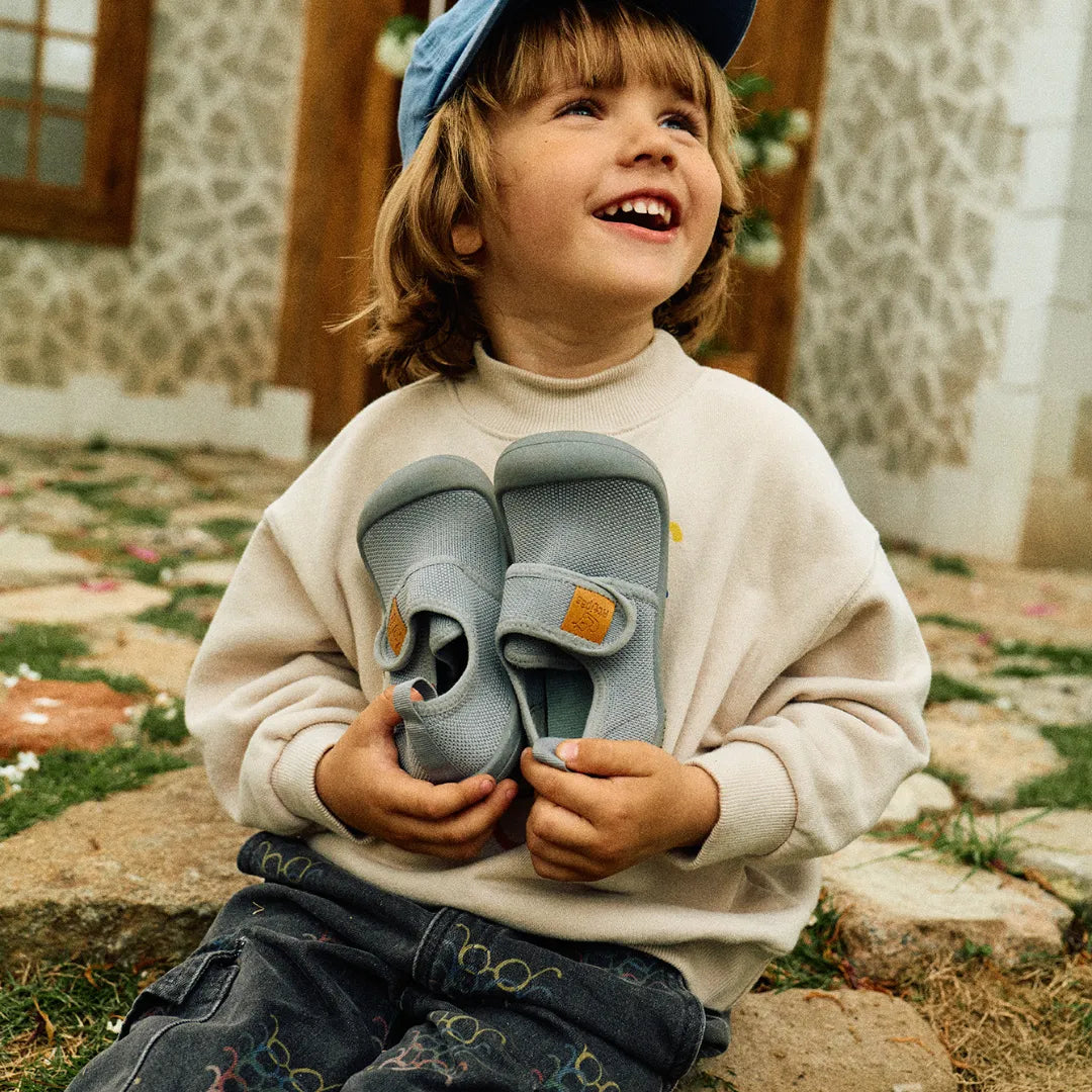 Menina sorridente segurando os Attipas Skin Shoes (Grey), transmitindo a leveza e bem-estar que os sapatos garantem no dia a dia.
