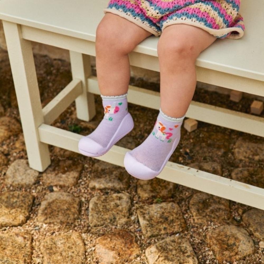 Bebé com calçado Attipas UNICORN PURPLE sentado num banco de madeira, mostrando a sola protetora em uso exterior sobre pedras.