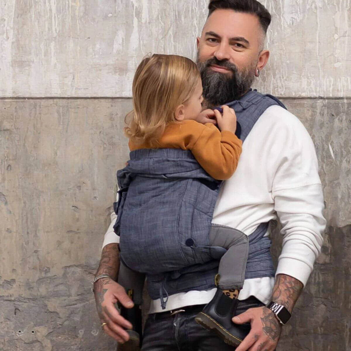 Pai carregando um bebé num Mei tai Hoppediz Híbrido Bondolino Plus Denim, mostrando o conforto e a proximidade entre pai e filho.