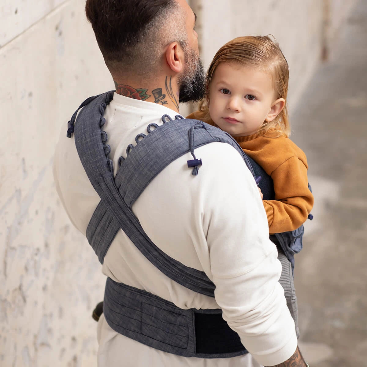 Vista traseira de um pai com um bebé num Mei tai Hoppediz Híbrido Bondolino Plus Denim, destacando a distribuição das alças e o suporte nas costas.