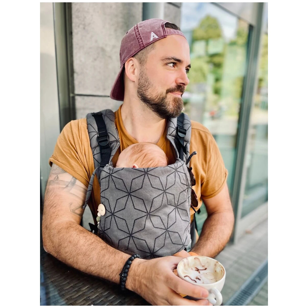 Homem sentado num café, usando a Mochila Hoppediz Ajustável BabySize Buckle Malmo Stone com um bebé, ilustrando a facilidade de uso e conforto do produto em situações do dia-a-dia.