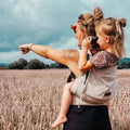 Mãe carregando seu filho num campo, usando a Mochila Hoppediz Ajustável Buckle Toddler Djerba, exemplificando o uso confortável e prático do produto.