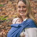 Mulher sorridente usando o pano elástico azul, com o bebé seguro e confortável na posição de babywearing.