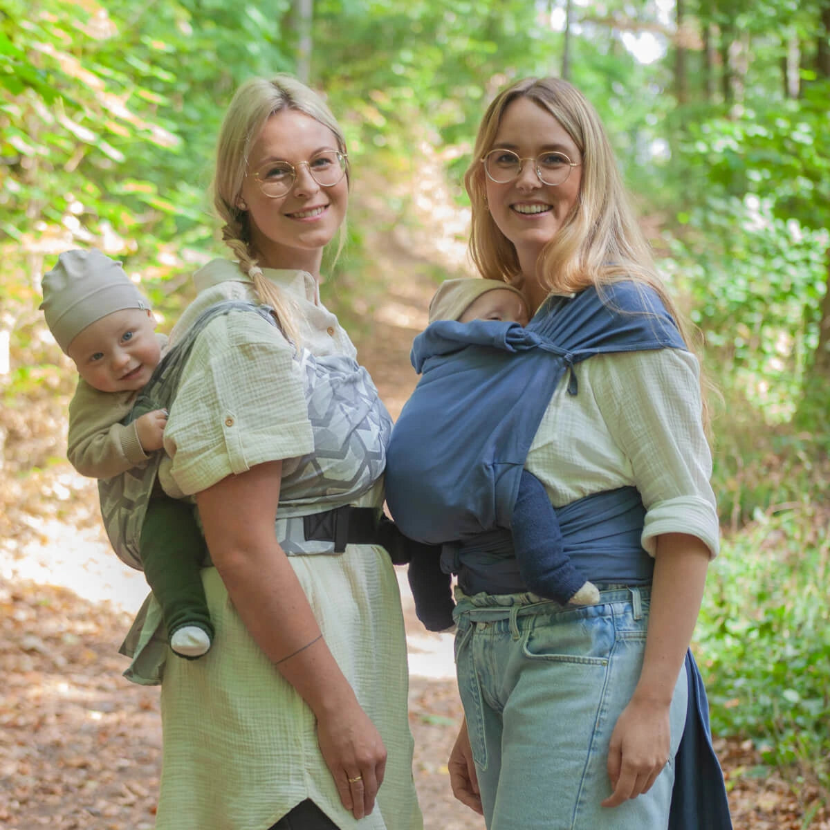 Duas mulheres usando panos elásticos azuis, cada uma carregando um bebé, caminhando numa trilha de floresta.