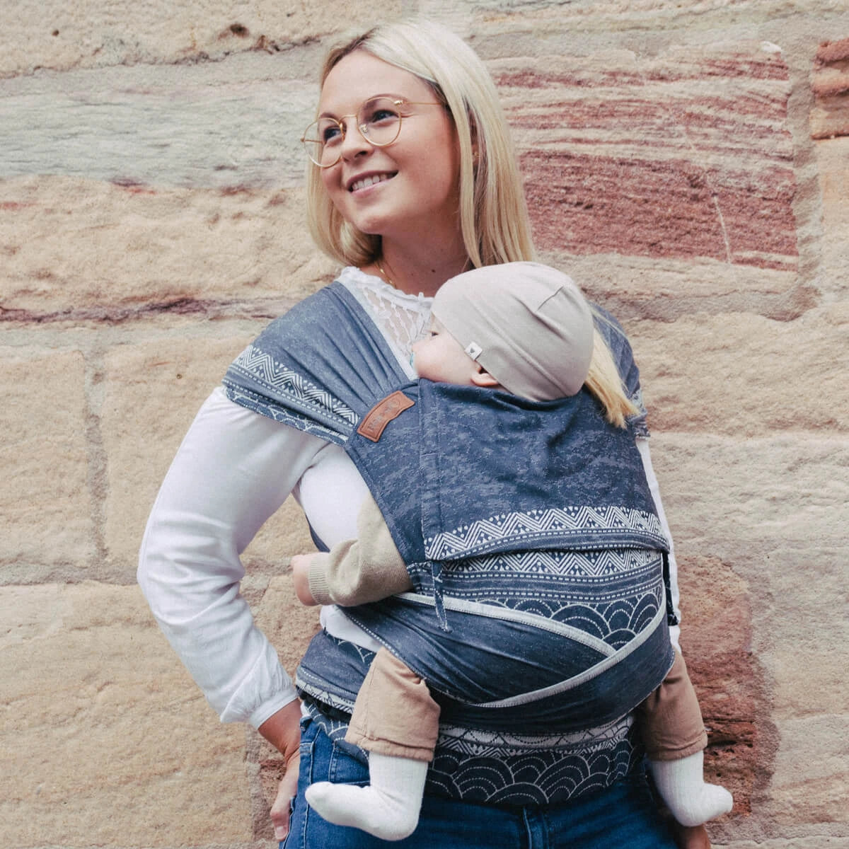 Mulher a usar o Mei Tai Hoppediz Evolutivo Hop Tye Advanced Sapporo, carregando um bebé na posição frontal, com um sorriso mostrando a facilidade de uso e o design elegante do porta-bebés.