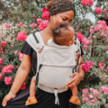 Mãe usando a Mochila Hoppediz Primeo Djerba com bebé na frente, em ambiente florido.