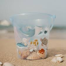 Balde de plástico azul claro com decorações de animais marinhos, cheio de conchas, pousado na areia da praia.