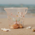 Balde de praia transparente Little Dutch com decoração de borboleta e flores, cheio de conchas na areia junto ao mar.