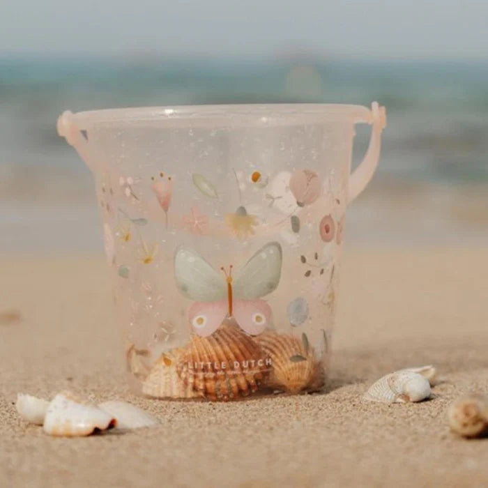 Balde de praia transparente Little Dutch com decoração de borboleta e flores, cheio de conchas na areia junto ao mar.