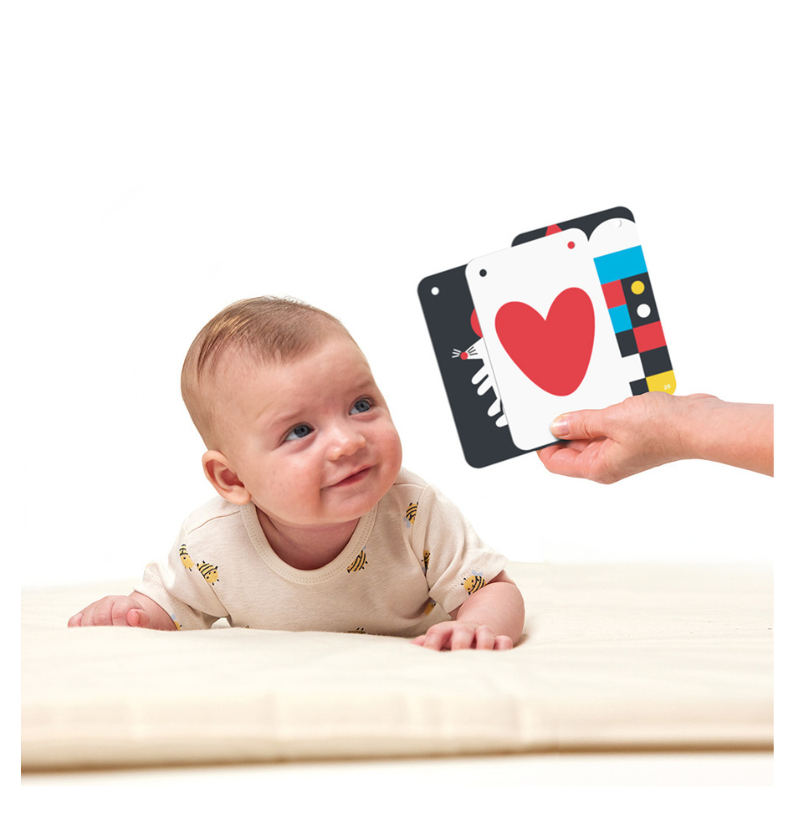 Um bebé deitado de barriga (tummy time) a olhar atentamente para um dos cartões de alto contraste que uma mão segura à sua frente.
