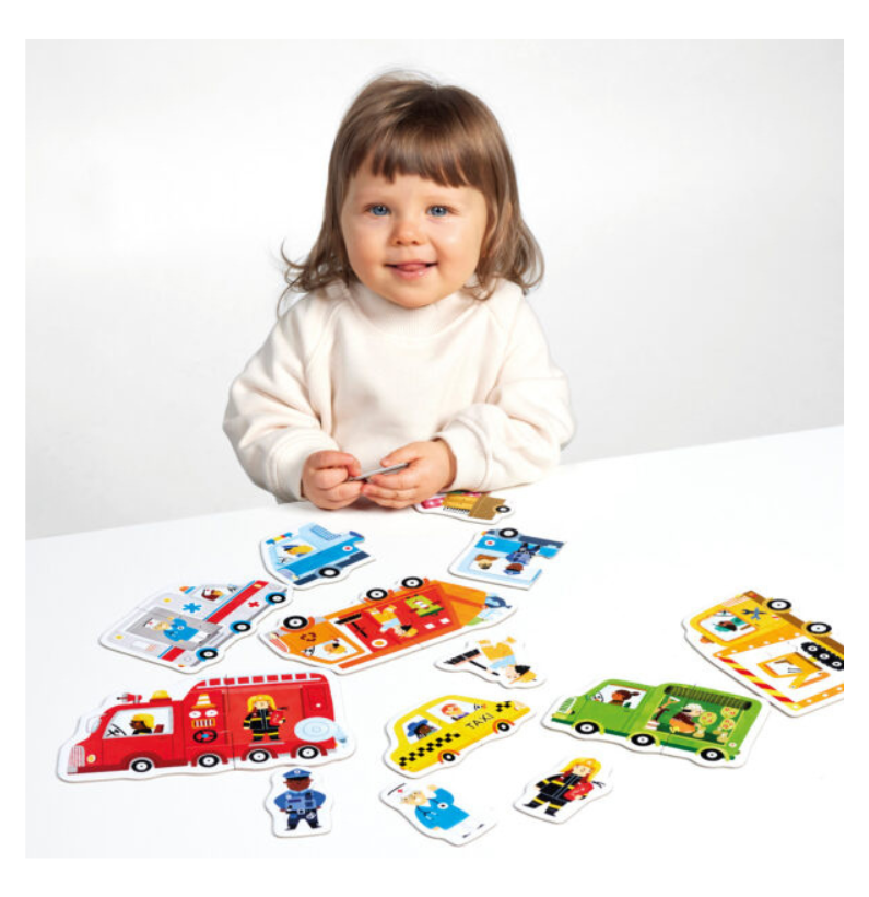 Uma criança pequena sorridente a brincar com as peças soltas dos puzzles de carros sobre uma mesa branca.