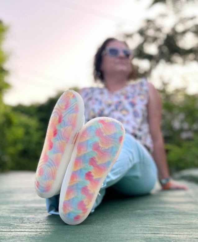 Pessoa sentada ao ar livre, usando o calçado barefoot adulto Mapis Arco Íris, destacando a sola colorida e o estilo casual.