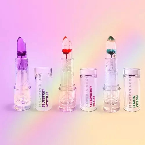 Três bálsamos labiais Inuwet Bálsamo de Karité "Flower in a Box" abertos, com flores roxa, vermelha e verde, e tampas correspondentes com aromas Mirtilo, Morango e Limão, sobre um fundo gradiente.