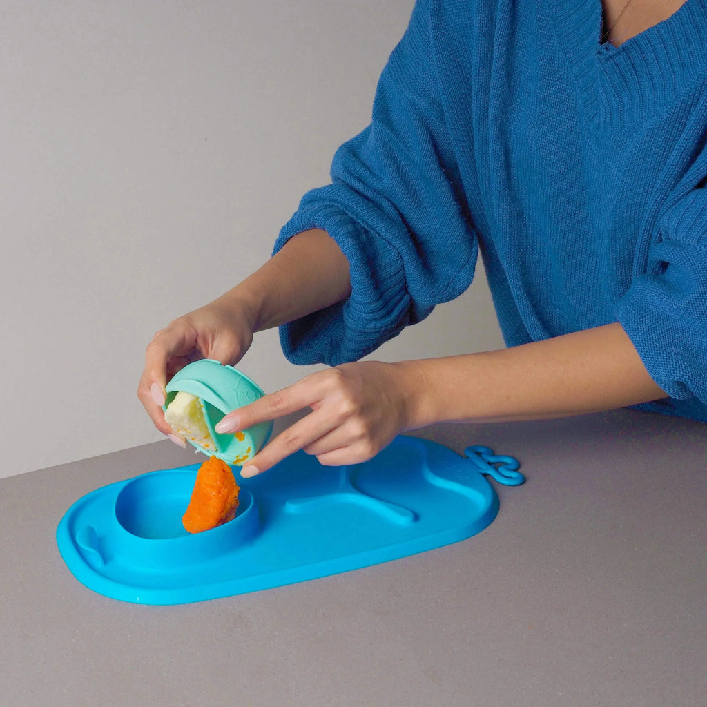 Mãos a servir puré de cenoura e outro alimento de uma taça b.box com divisória para um prato de bebé.