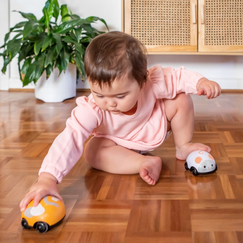 Bebé a gatinhar no chão de madeira, a interagir com os brinquedos Pull Backs Gato (laranja) e Rato (branco).