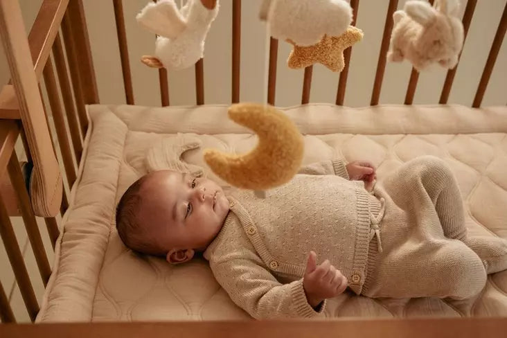 Bebé deitado pacificamente no berço, com a lua de peluche do móbile a pairar suavemente por cima.