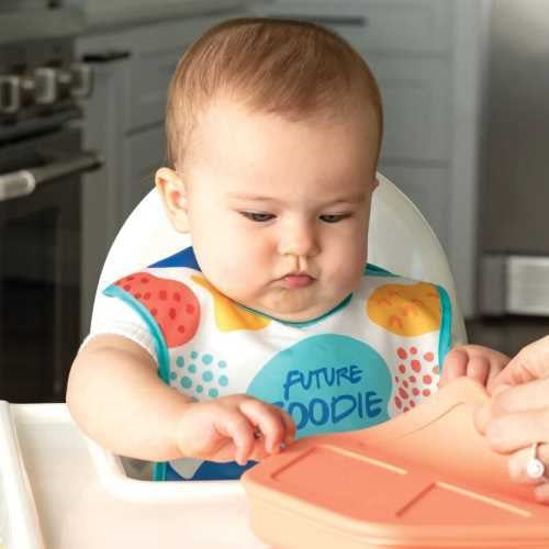 Bebé sentado numa cadeira alta a usar o babete colorido, a interagir com um recipiente de comida de silicone.