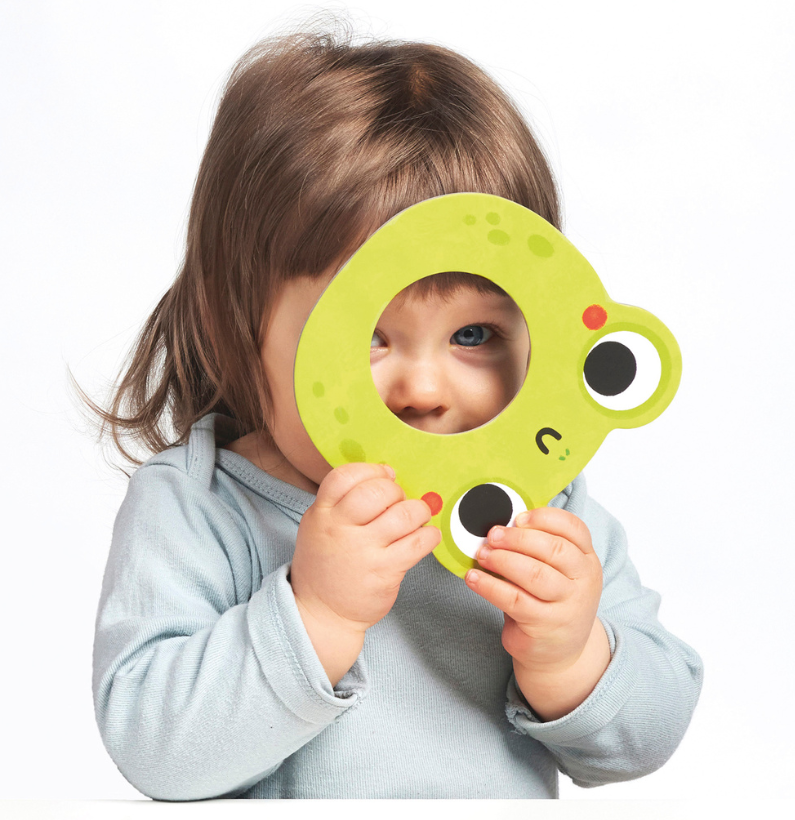 Bebé a brincar com uma peça do puzzle, segurando a parte maior do sapo verde que cobre parcialmente o seu rosto.