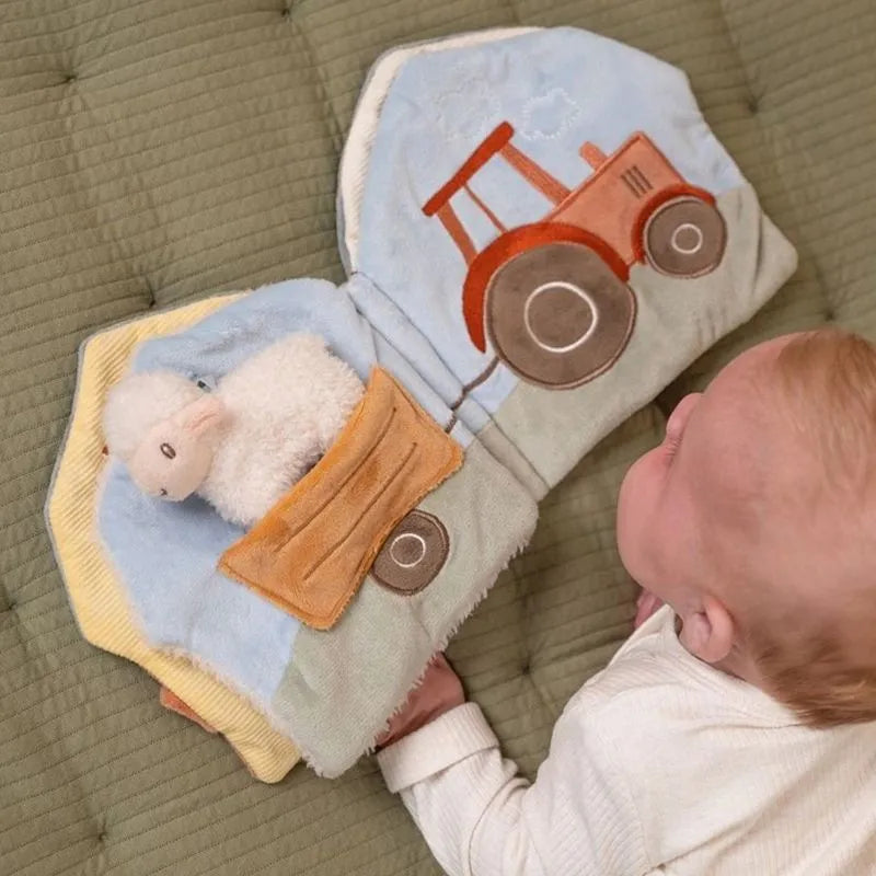 Bebé a segurar e a interagir com um livro de atividades de tecido aberto, que mostra um trator e uma ovelha de peluche.