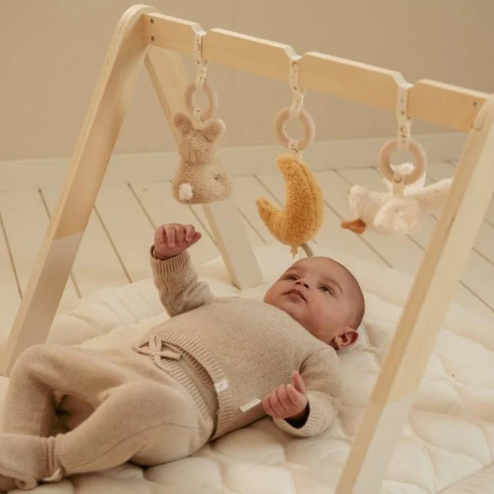 Bebé deitado de costas no chão, a olhar para cima para os brinquedos do ginásio de atividades Little Dutch. O bebé veste roupa de malha bege.