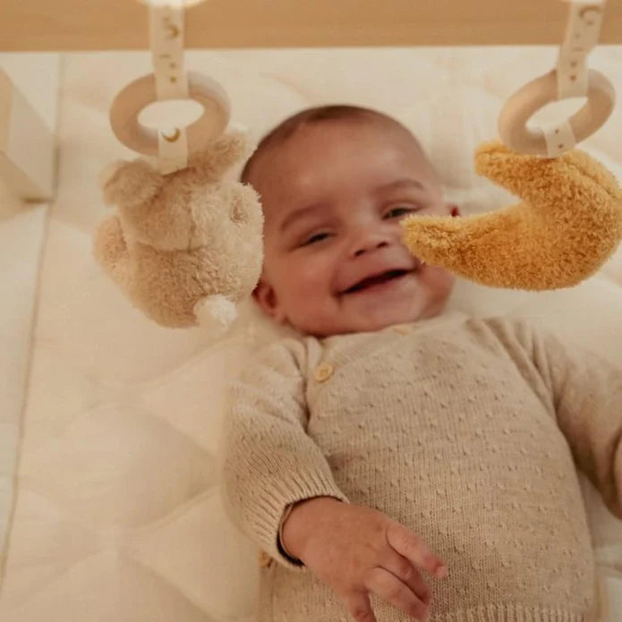 Close-up de um bebé sorridente deitado de costas, olhando para os brinquedos pendurados do seu ginásio de atividades Little Dutch.