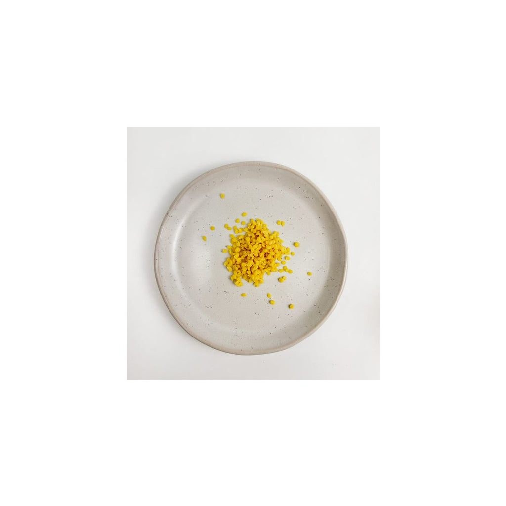 Pérolas de cera de abelhas orgânica amarela natural, da marca Gotu, dispostas num pequeno monte sobre um prato de cerâmica cinza salpicado.