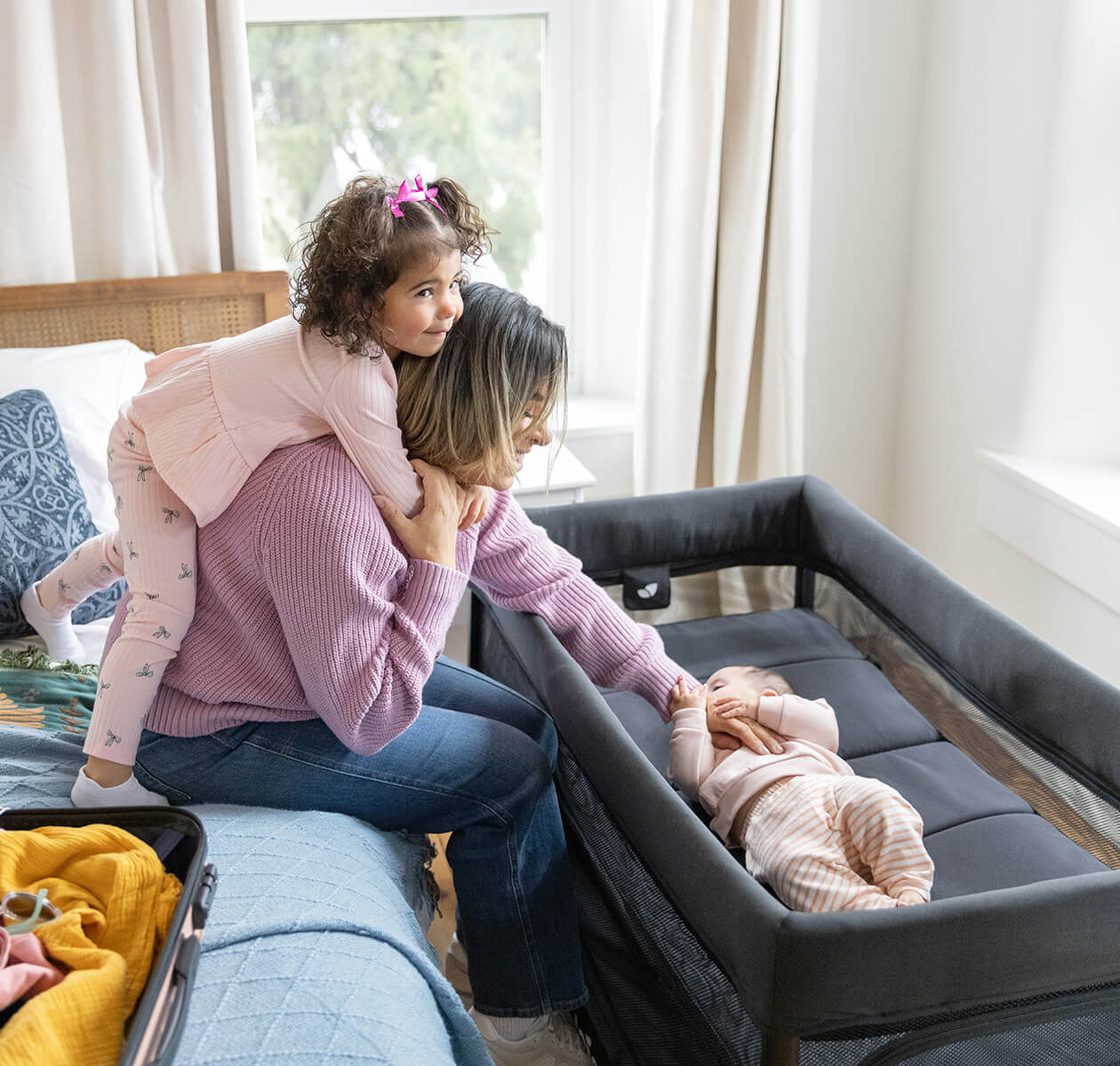 Mãe sentada na cama, com uma criança ao pescoço, a interagir com um bebé que está deitado no moisés do berço de viagem Joie amigo™ 120.