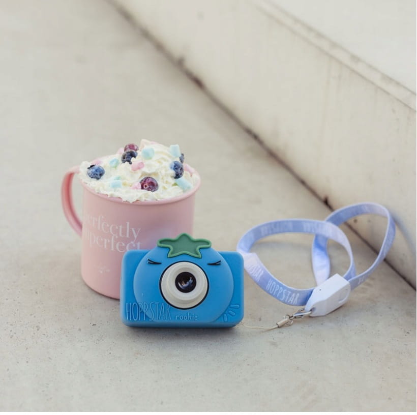 Máquina fotográfica Hoppstar Rookie Bluebelle ao lado de uma caneca rosa, mostrando o cordão e o design divertido do produto.