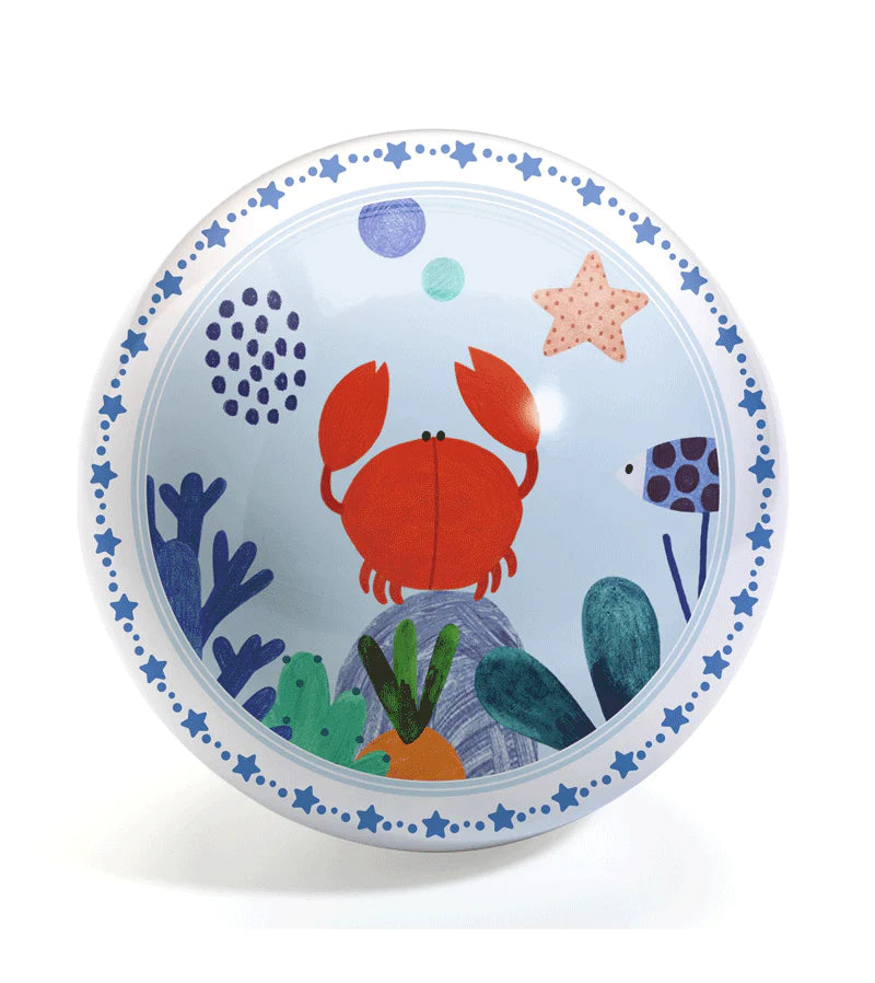 Bola de brincar redonda com ilustração central de um caranguejo vermelho rodeado por algas e estrelas do mar num fundo azul claro. A borda tem um padrão de estrelas azuis.
