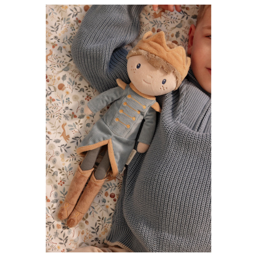 O boneco Príncipe Jim deitado ao lado de uma criança que dorme ou descansa, sobre um lençol com padrão floral e animal.