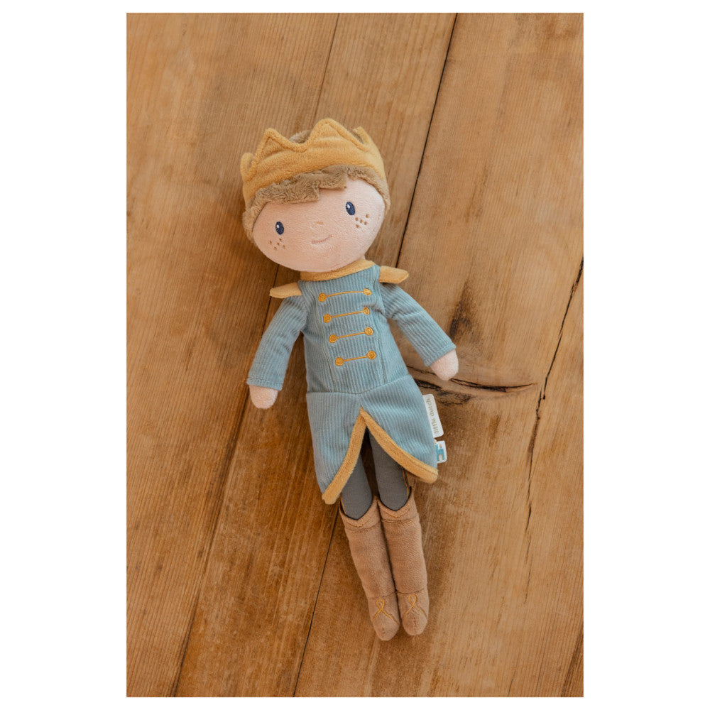 Boneco de peluche Príncipe Jim da Little Dutch com coroa dourada e casaco azul, deitado sobre um soalho de madeira.