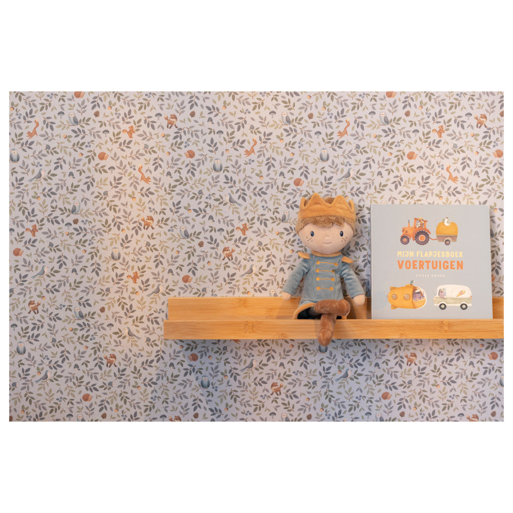 Boneco Príncipe Jim sentado numa prateleira de madeira, ao lado de um livro infantil com motivos de veículos, contra uma parede com papel de parede de folhagem.