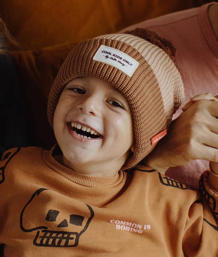 Close-up de criança sorridente a usar o gorro HELLO HOSSY com pompom Coffee, destacando o rótulo.