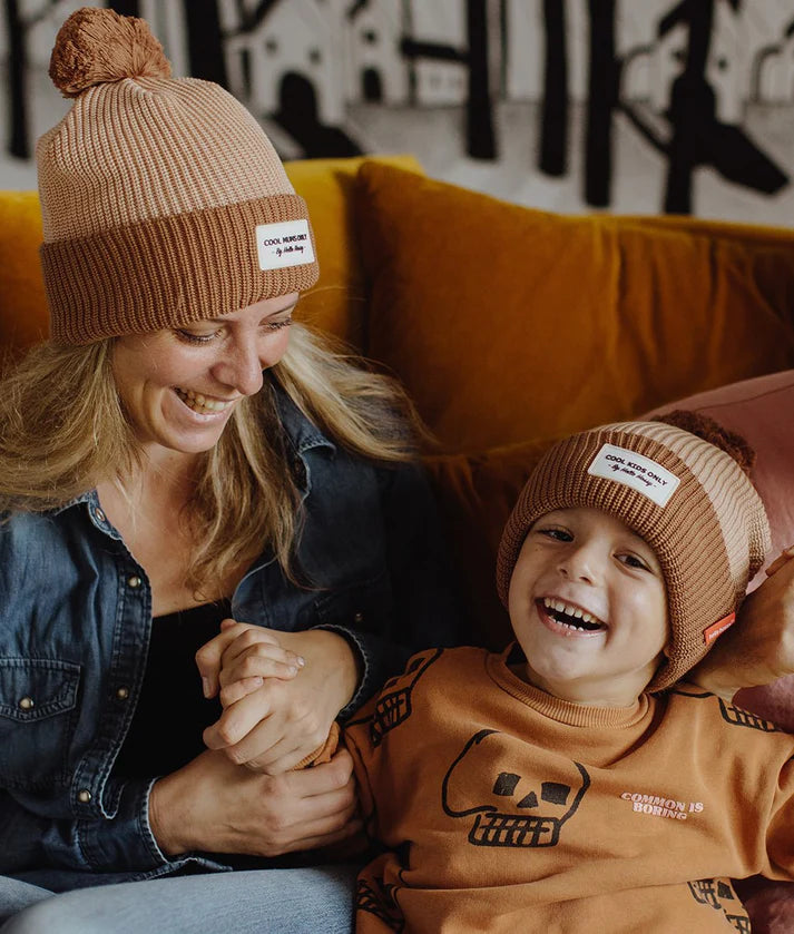 Mãe e filho sorridentes, ambos com o gorro HELLO HOSSY com pompom Coffee, abraçados num sofá.