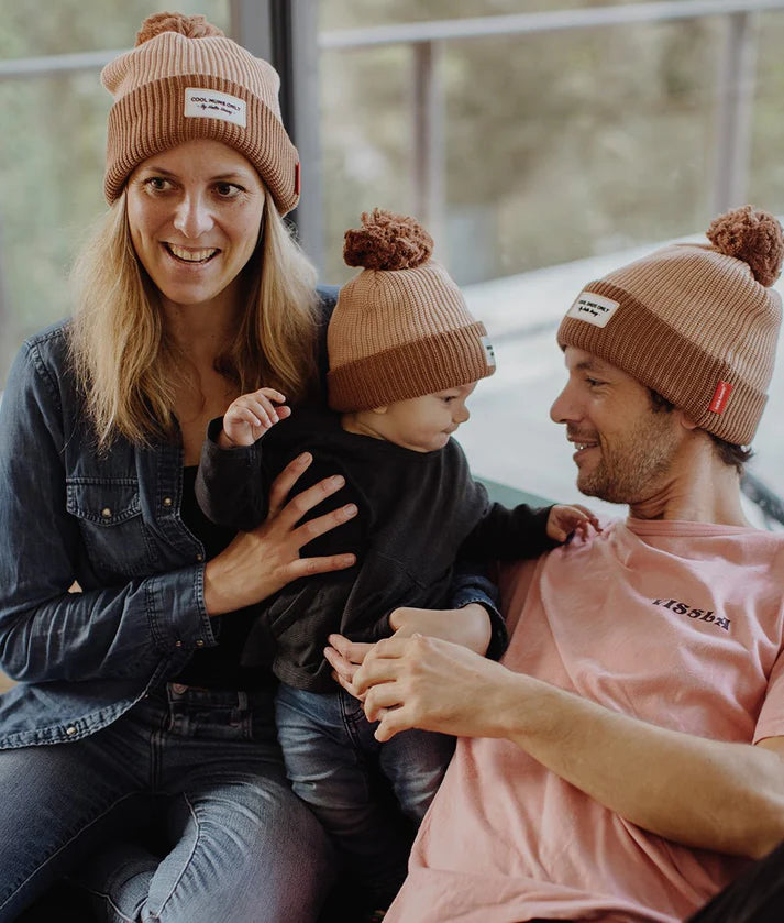 Família (pai, mãe e bebé) com os gorros HELLO HOSSY com pompom Coffee, num ambiente acolhedor.