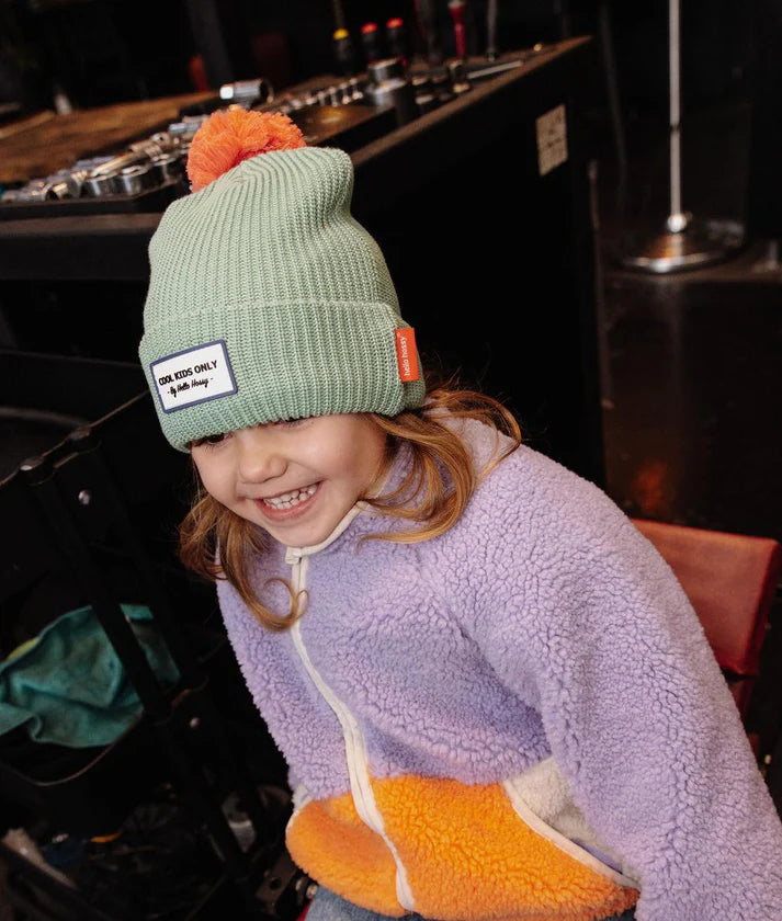 Menina com gorro Hello Hossy verde-menta com pompom laranja, num ambiente de oficina.